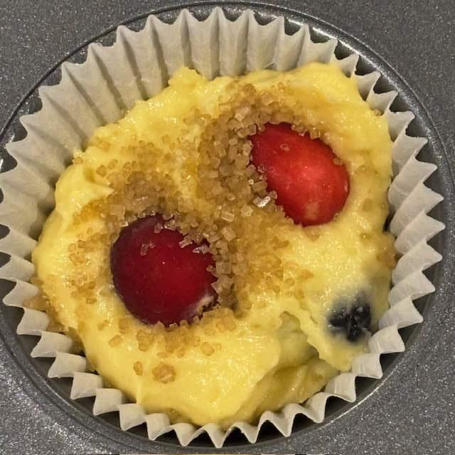 Cake mix muffins batter in a paper muffin cup with cranberries and turbinado sugar on top of the batter.