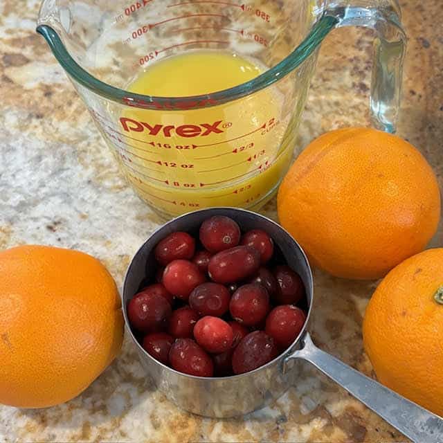 Cup of orange juice, cup of cranberries and 3 oranges on a kitchen counter.