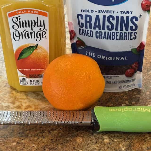 Orange juice, dried cranberries, orange and zester tool on kitchen counter.