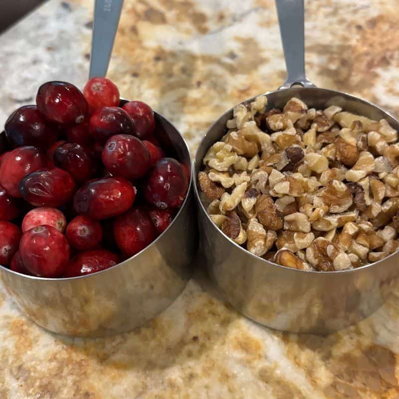 A cup of cranberries and a cup of chopped walnuts on a kitchen counter.